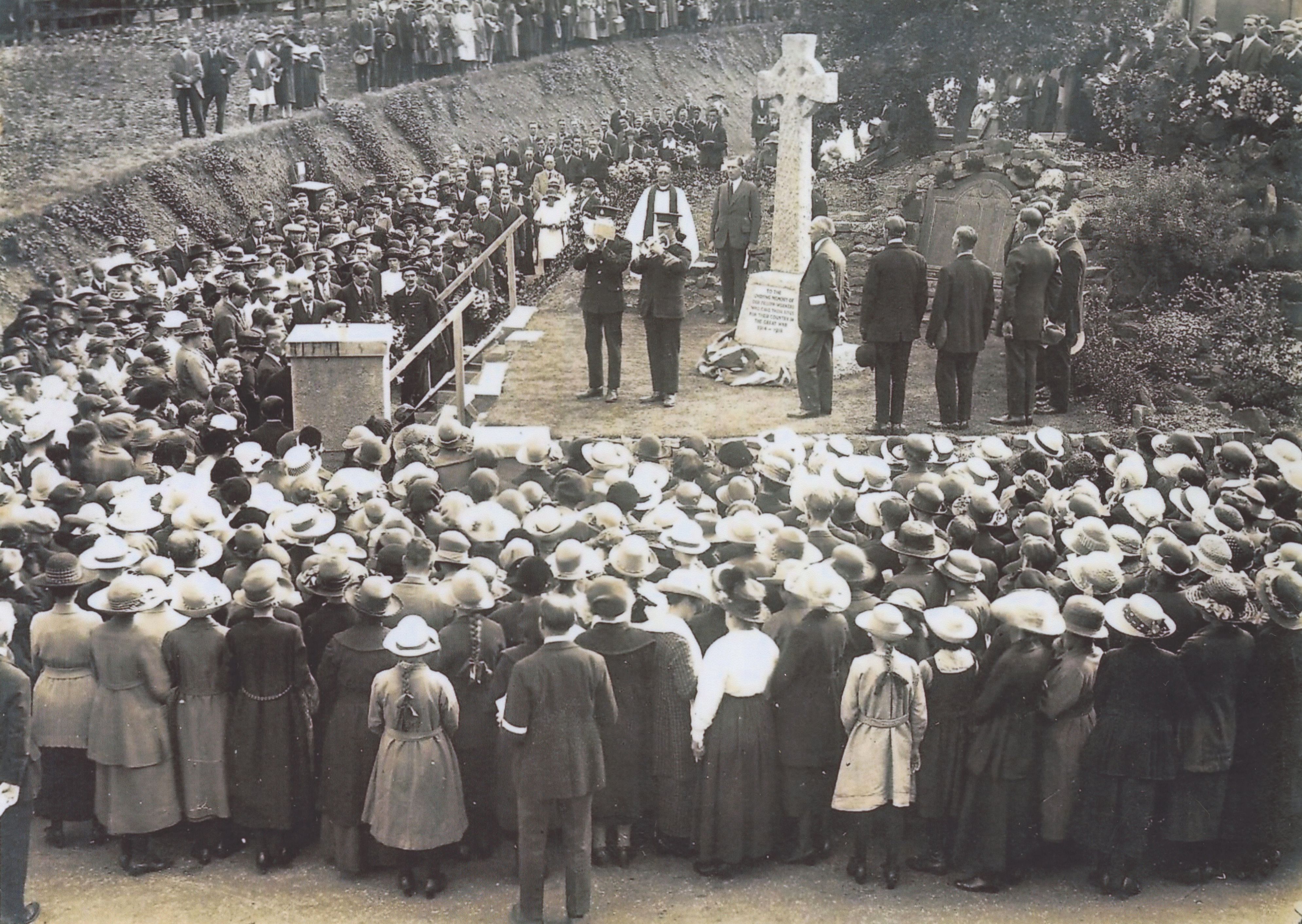 A sepia photograph of a service at the Apsley War Memorial. The Managing Director, Mr Ling, stands in the centre before the memorial's cross. There are two groups stood either side of the memorial, and a crowd watches on. 