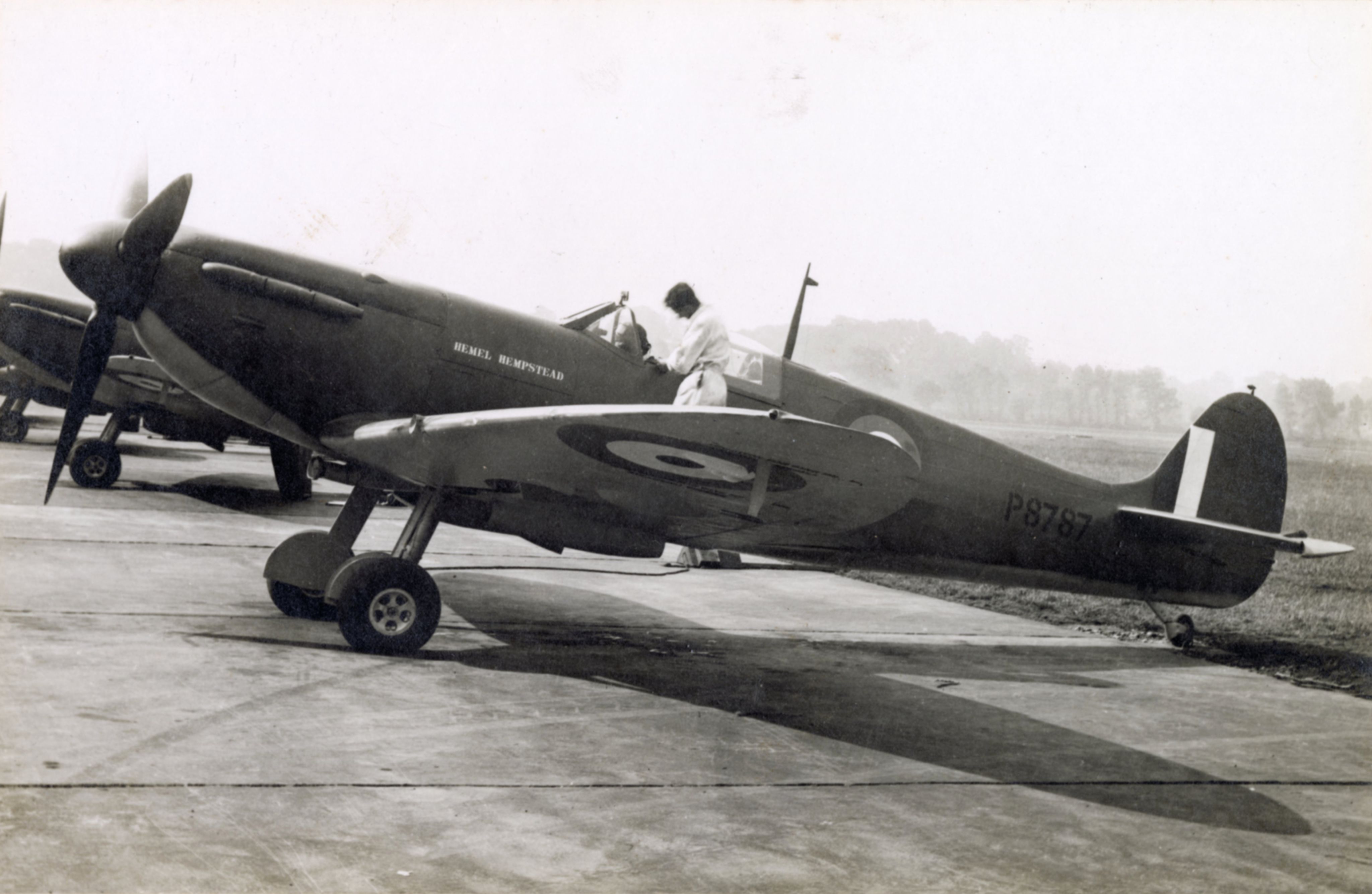 A black and white photograph of a Spitfire plane bearing the name 'Hemel Hempstead'. A man stands beside the cockpit.