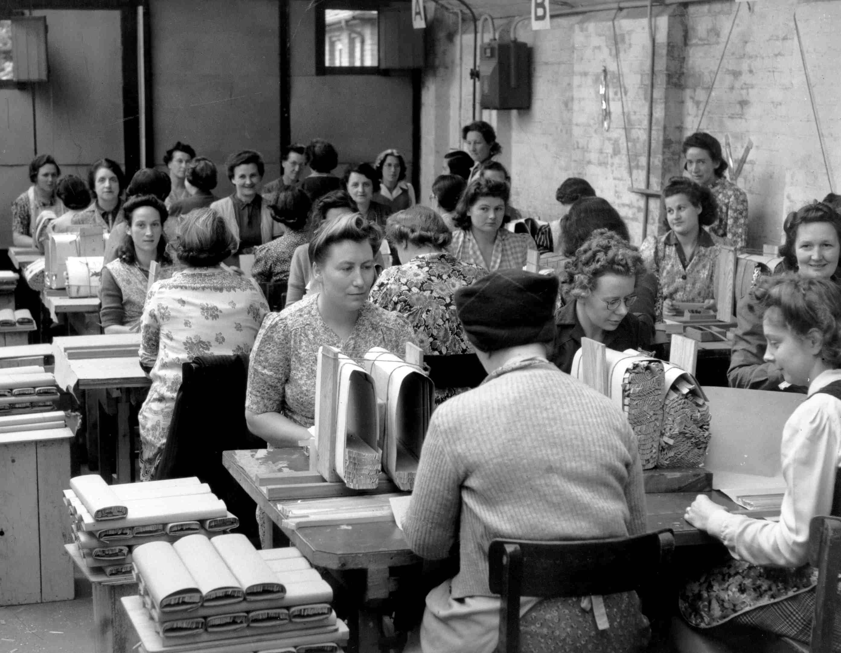 A black and white photo of a group of women sat at rows of tables, packaging 'window' foil strips. 