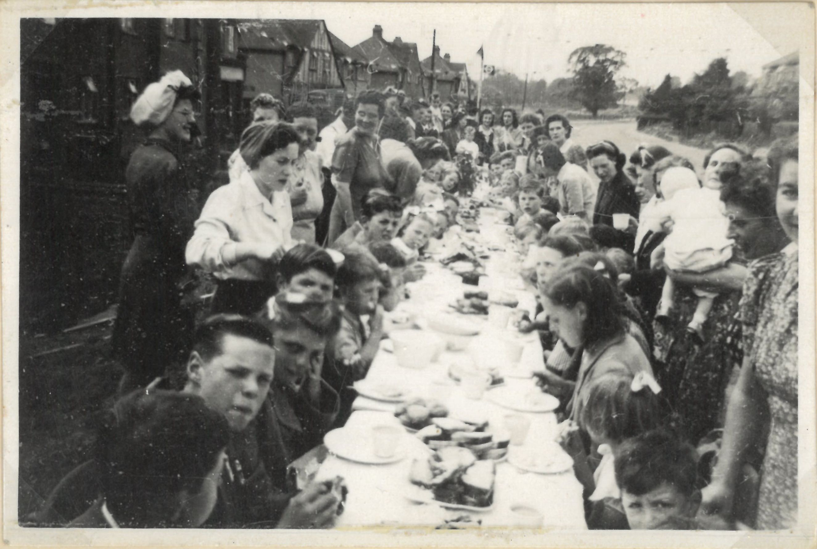 A black and white photograph depicting a street party. A long table with plates of food is at the centre of the image, with many children and adults sat or stood at either side. 