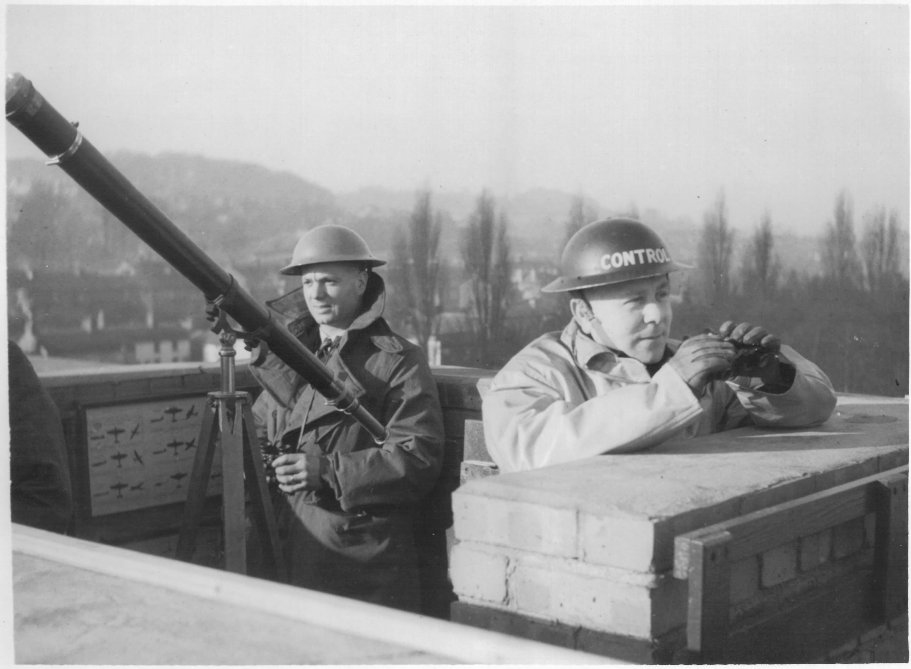 A black and white photograph of two men stationed on a rooftop. Both are wearing metal helmets and holding binoculars. The man on the left is stood next to a telescope. A chart of aircraft shapes is in the background. 