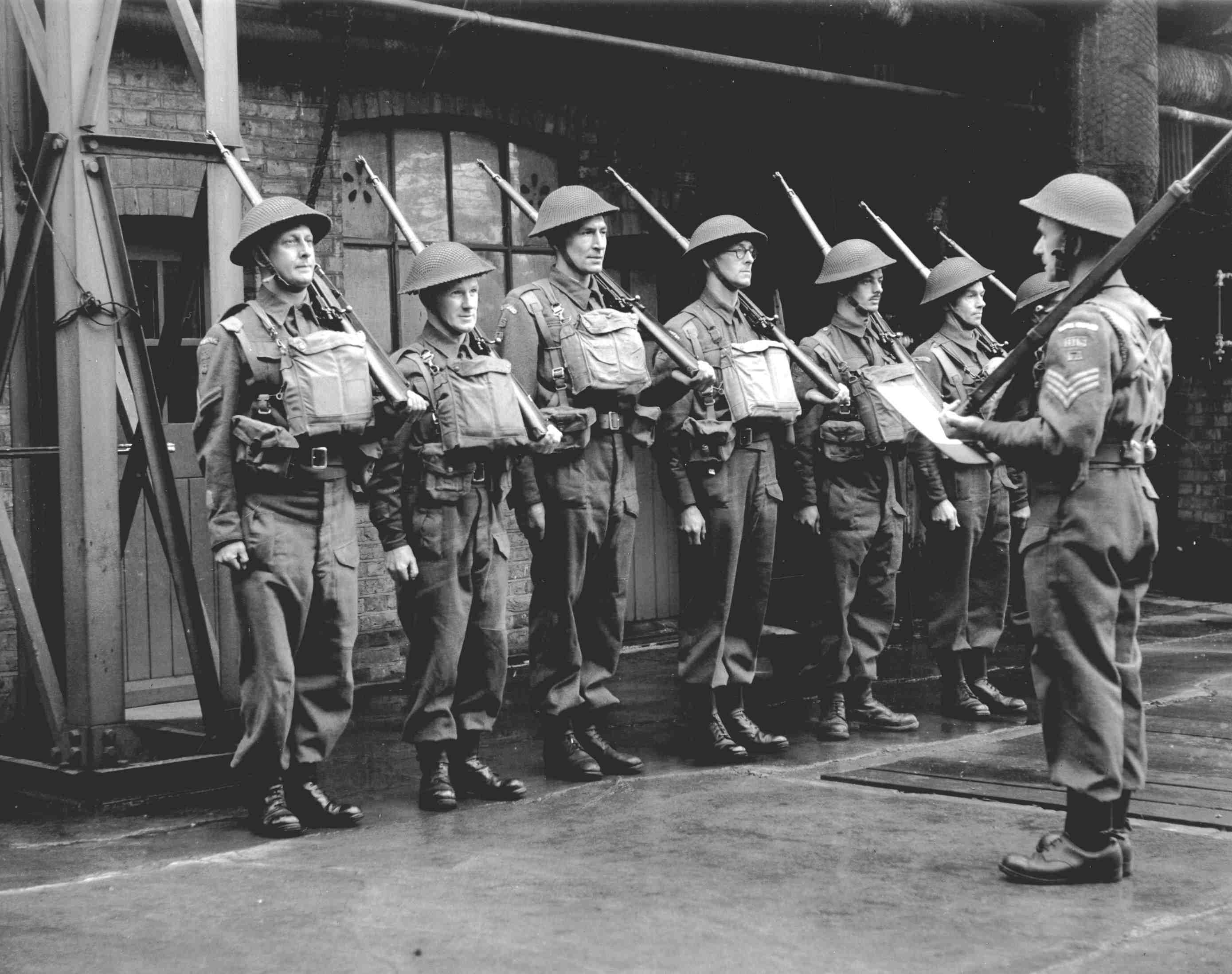 A black and white photograph showing six men wearing army uniforms lined up and with guns on their shoulders. Their officer, also in uniform, stands in front with his back to the camera.
