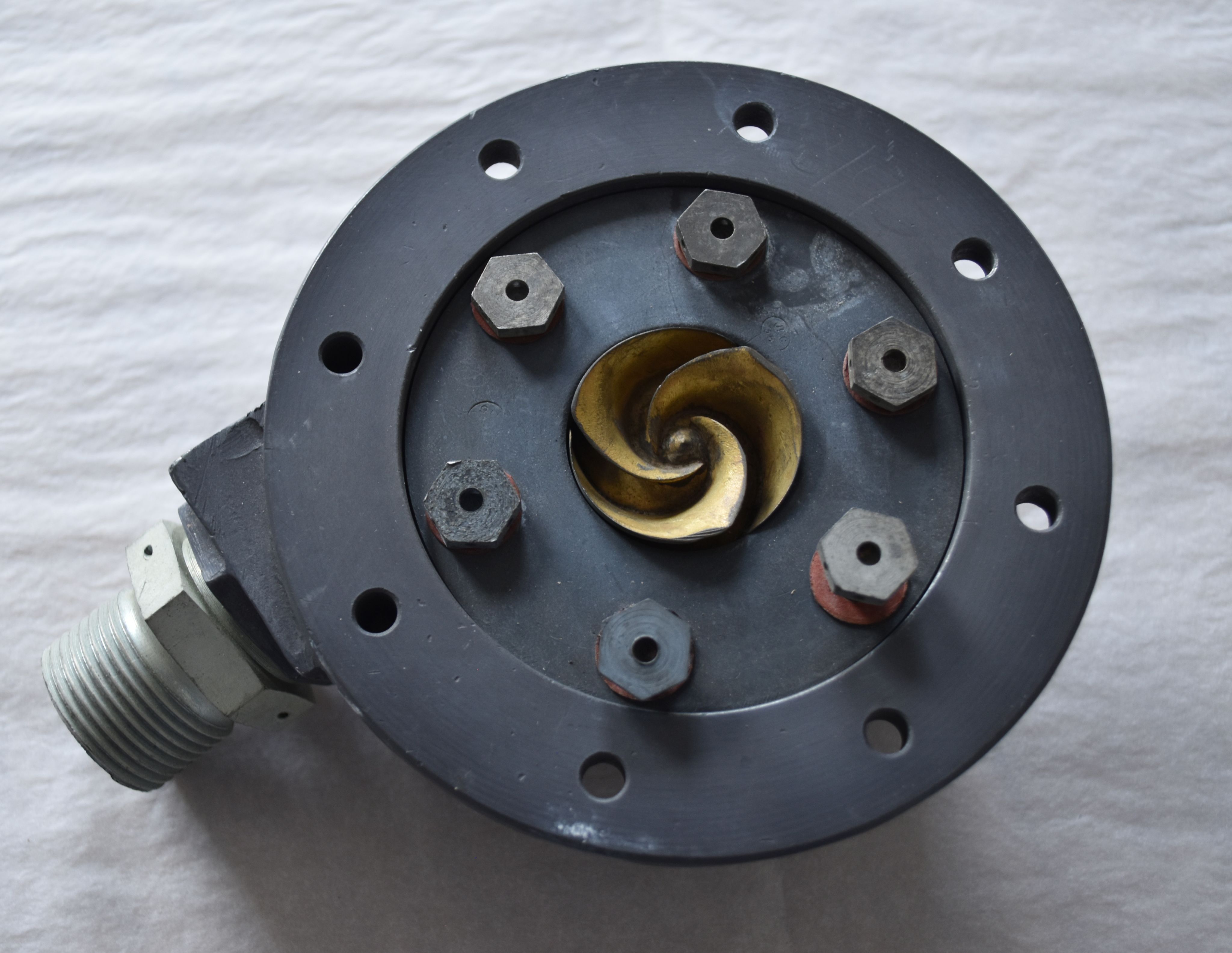 A liquid fuel pump. A black circular object with central turbine and six central bolts, and a screw nozzle in the lower left.